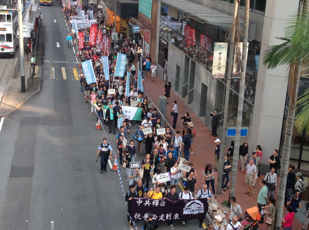 Eine große Gruppe von Menschen marschiert auf einer Demonstration durch eine Stadtstraße, hält Schilder und Plakate hoch, mit Bäumen, Glasgebäuden und Fahrzeugen auf den Seiten.