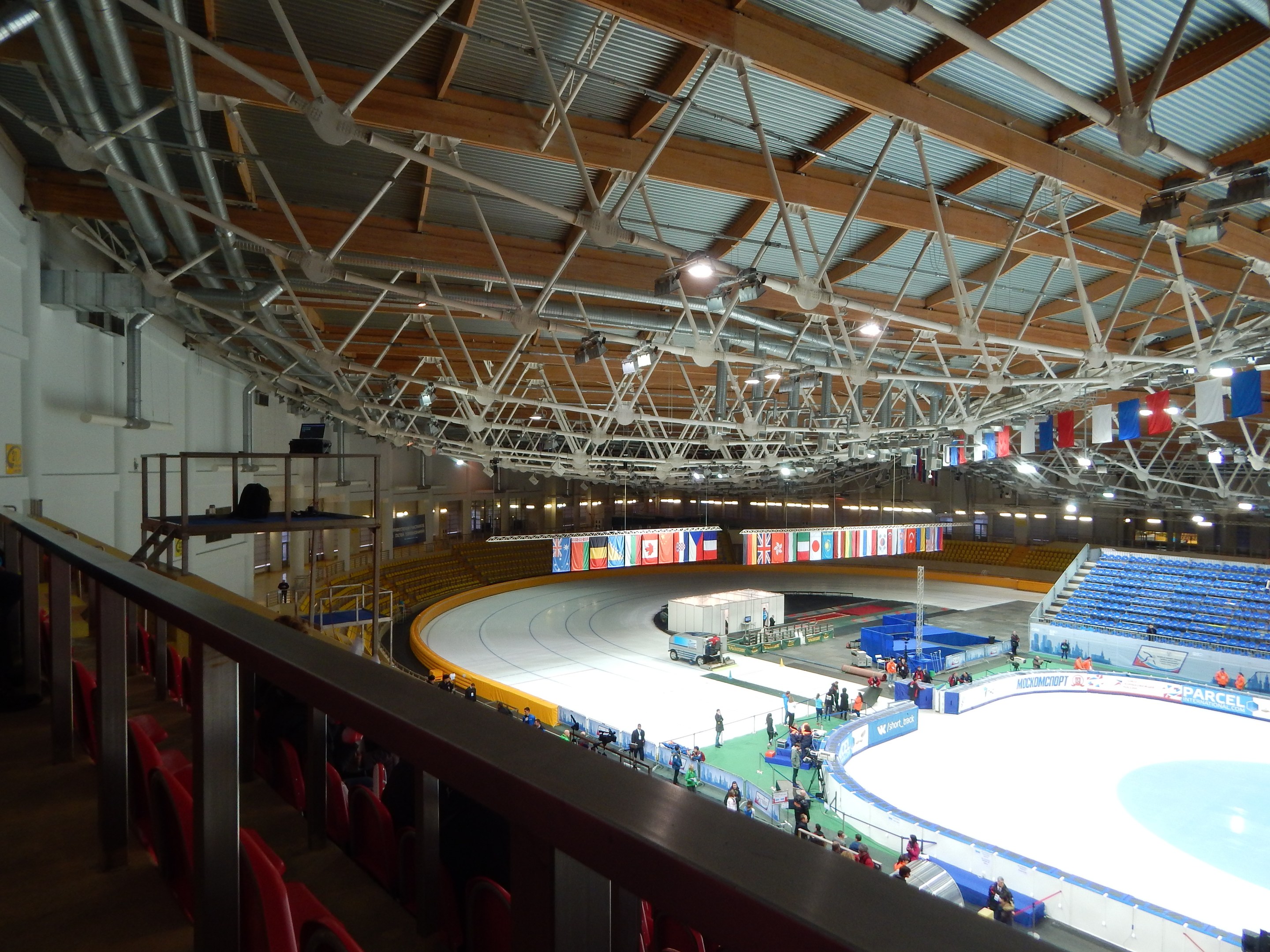 Große Indoor-Eisbahn voller Menschen, umgeben von Sitzplätzen, Geländern, Brettern, Beleuchtungskörpern und strukturellen Elementen, mit einer Wand im Hintergrund.