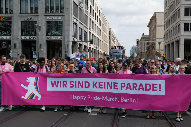Eine Gruppe von Menschen geht eine Straße in Berlin, Deutschland, entlang und hält eine pinkfarbene Fahne mit der Aufschrift 'Happy Pride March'. Die Straße ist von Gebäuden mit Fenstern, Laternenmasten und Verkehrssignalen gesäumt.