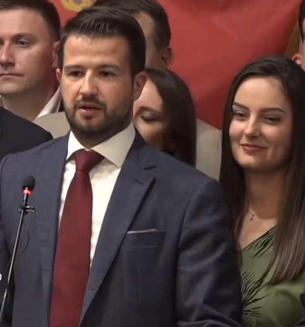 Ein Mann in Anzug und Krawatte spricht in ein Mikrofon an einem Rednerpult mit einem Banner im Hintergrund zu einer Menge.