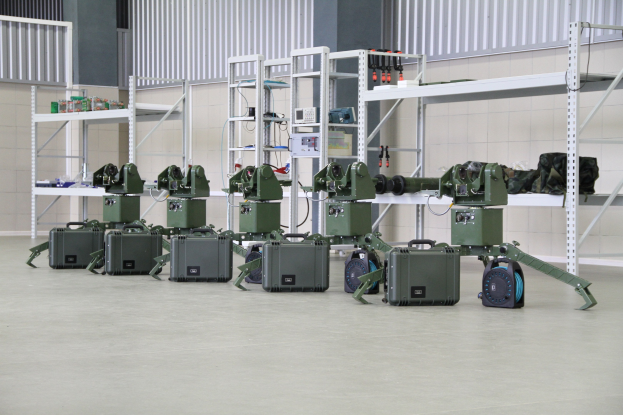 Gruppe von Militärrobotern auf einem Boden mit Koffern und Objekten auf Gestellen, vor einer Wand mit Geländer.