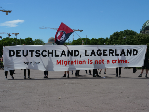 Eine Gruppe von Menschen mit einem Transparent 'Deutschland, Lagerland Migration ist kein Verbrechen' und einer Flagge, mit Straßenlaternen, Bäumen, Kränen, einem Gebäude und einem bewölkten Himmel im Hintergrund.