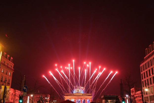 Eine nächtliche Stadtstraße in Berlin mit dem Brandenburger Tor in der Mitte, umgeben von beleuchteten Gebäuden, Bäumen, Pfosten, Verkehrszeichen, Schildern und Flaggen, mit Feuerwerk, das den Himmel über dem Tor erhellt.
