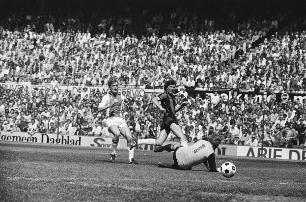 Schwarzes und weißes Foto von Männern, die auf einem Feld Fussball spielen, mit Zuschauern im Hintergrund und einer "Bundesliga 1978-79 Ajax 1-1 Hannover 96"-Banner.