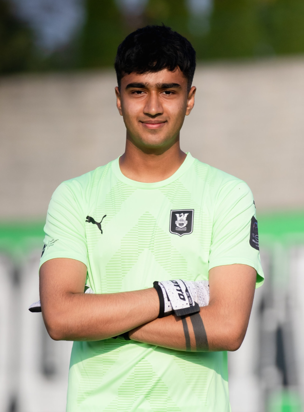 Ein junger Mann in einem grünen T-Shirt steht auf einem Fußballfeld und hat die Arme verschränkt, während er lächelt und der Hintergrund unscharf ist.