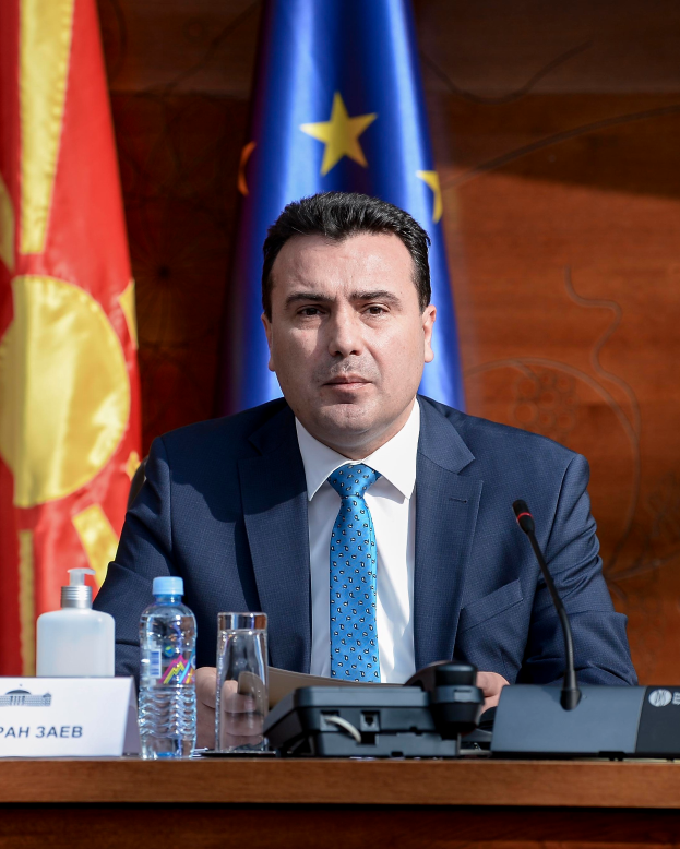 Ein Mann im Anzug und Krawatte sitzt an einem Tisch mit einem Mikrofon, Flaschen, einem Glas und einer Namensschilder, mit Flaggen im Hintergrund.
