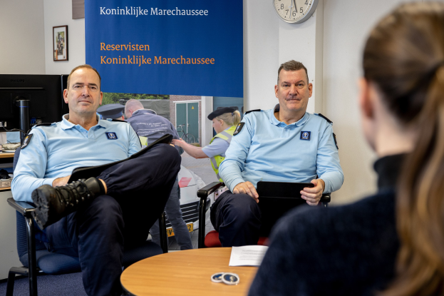 Zwei Polizeibeamte sitzen an einem Tisch mit einer Frau, umgeben von Papieren und Gegenständen, mit einem Wandbanner, einer Uhr und Fotorahmen im Hintergrund und einem zweiten Tisch mit einem Monitor, Tastatur und Maus links daneben.