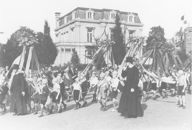 Schwarz-weißes Foto einer Gruppe von Menschen, die eine Straße entlanggehen und Fahnen schwenken, mit Hüten, einem Maibaum, Bäumen, Gebäuden und Himmel im Hintergrund.