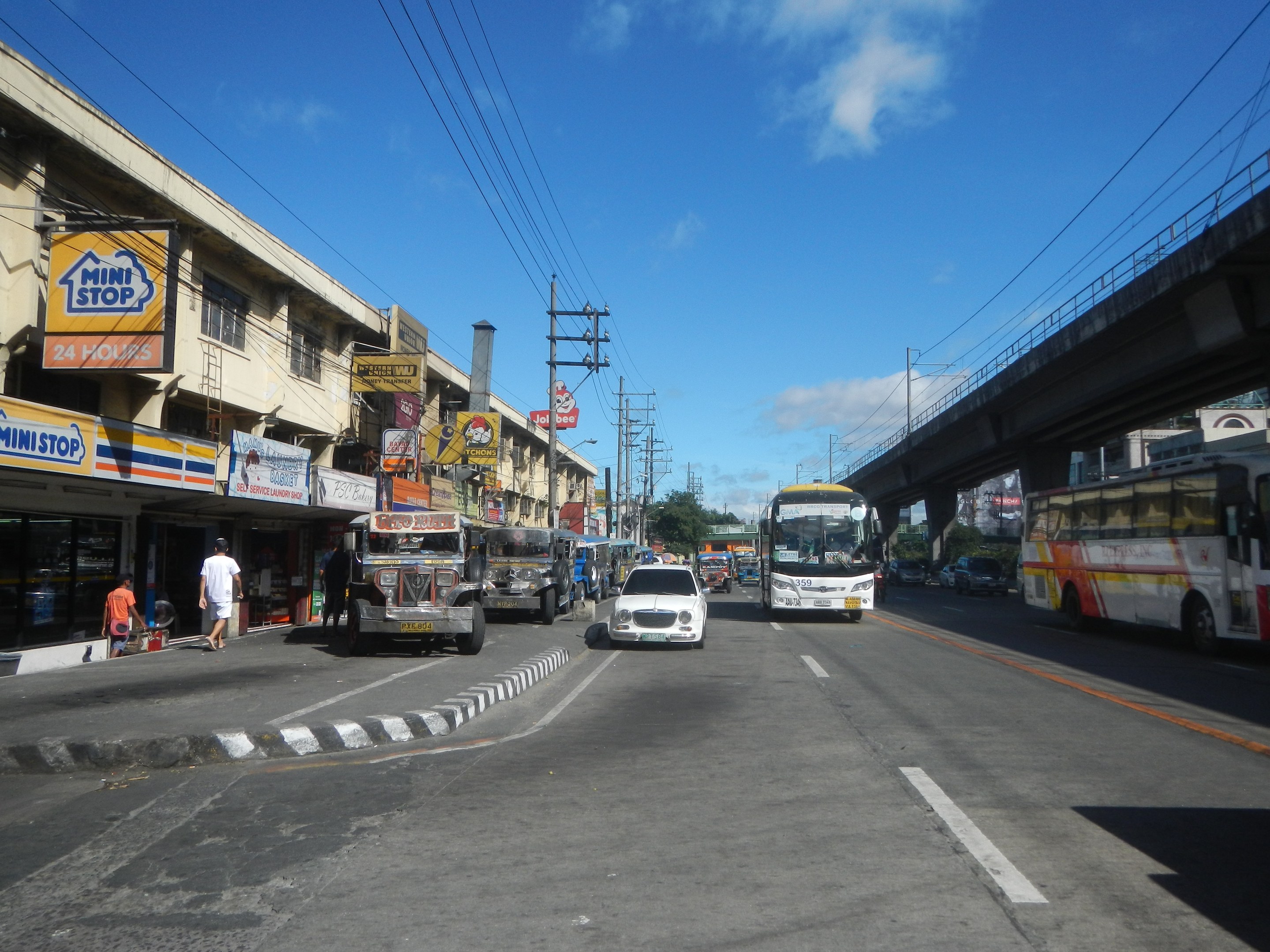 Volles Stadtbild mit Autos, Bussen, Fußgängern, Gebäuden, Strommasten, einer Brücke, Bäumen und einem bewölkten Himmel.