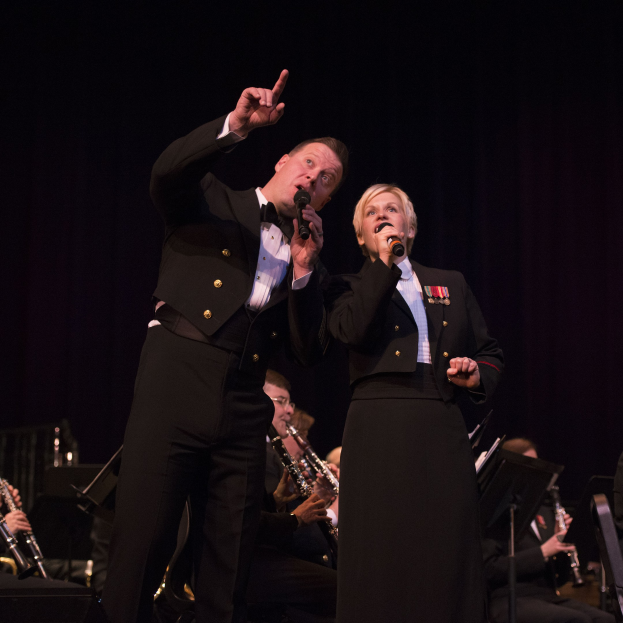 Ein Mann und eine Frau in Smokings, die Mikrofone auf einer Bühne halten, mit einer Sitzreihe von Musikern dahinter in einer dunklen Umgebung.