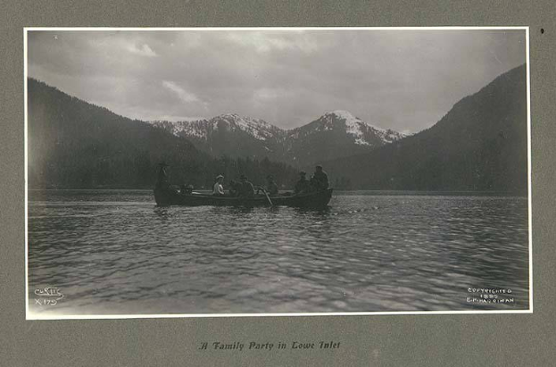 Schwarz-weiß-Foto einer Familie in einem Kanu auf einem See, umgeben von Bergen und Bäumen, mit Himmel im Hintergrund und Text unten.