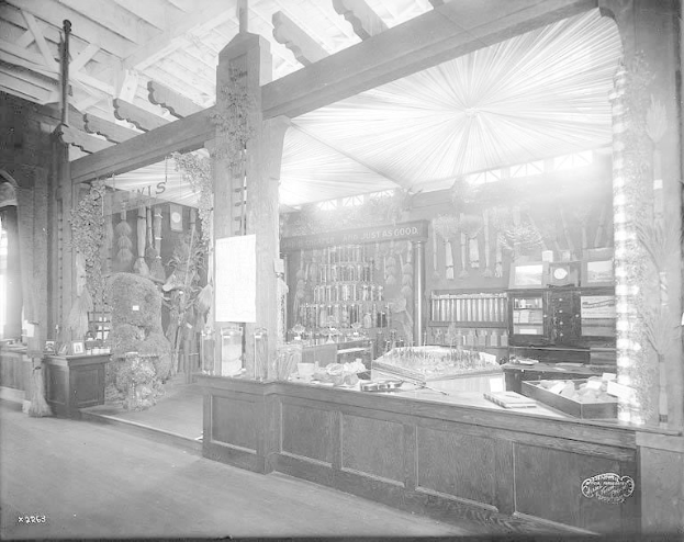 Schwarz-weiß-Foto eines alten Apothekenladens mit einem zentralen Tresen, auf dem verschiedene Gegenstände ausgebreitet sind, Regalen im Hintergrund gefüllt mit Flaschen und Artikeln und Text unten, der darauf hinweist, dass es sich um eine Apotheke handelt.