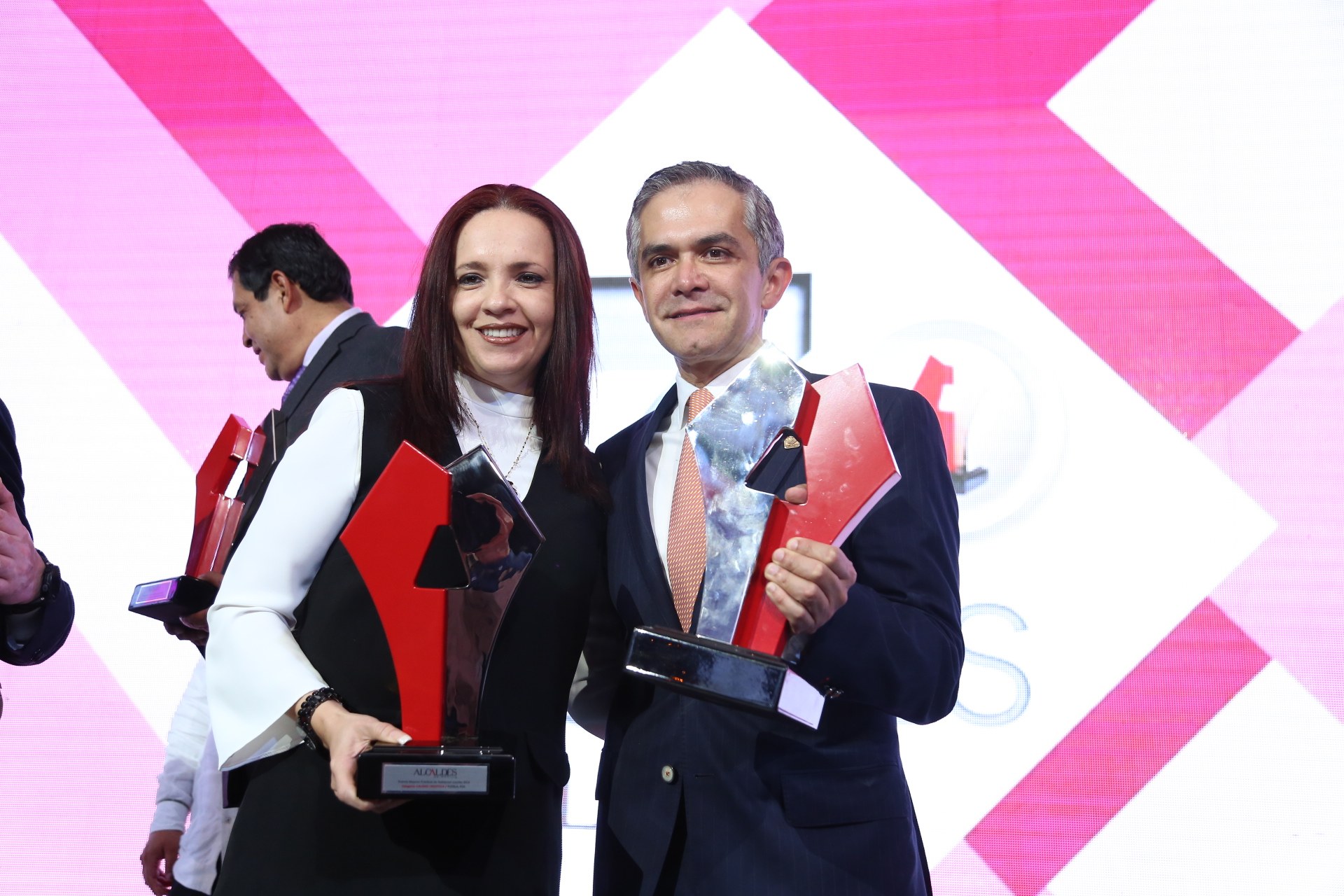 Zwei Menschen, die strahlend lächeln und Trophäen halten, auf einer Bühne mit einem Bildschirm im Hintergrund.