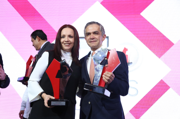 Zwei Menschen, die strahlend lächeln und Trophäen halten, auf einer Bühne mit einem Bildschirm im Hintergrund.