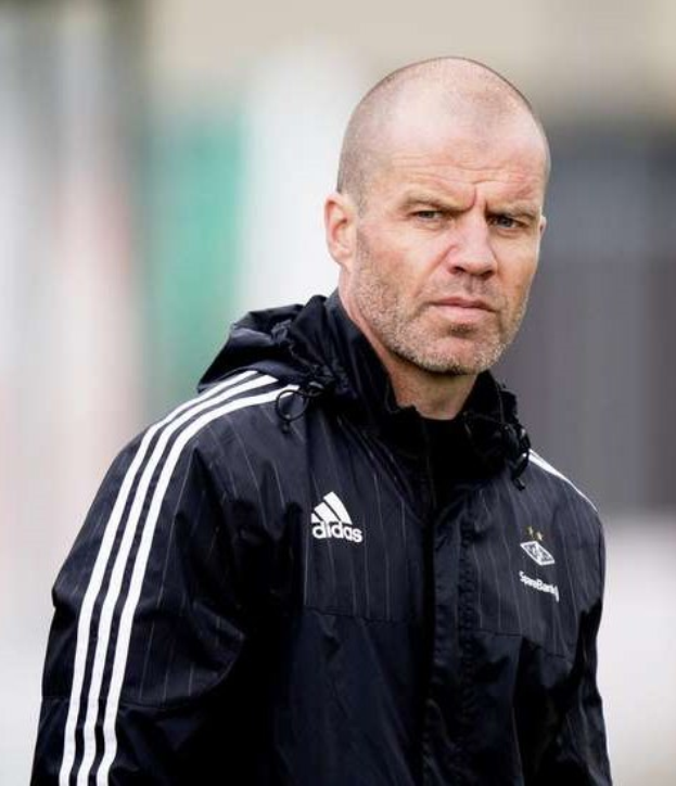 Baldiger Trainer in einer blauen Jacke mit weißen Streifen und weißem Kragen, der auf einem Fußballfeld steht, mit verschränkten Händen und ernster Miene, unscharfer Hintergrund.