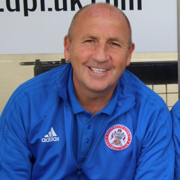 Zwei Männer mit einem blauen Jackett und einem Mann ohne Jackett sitzen nebeneinander vor einer Wand mit einer Tafel im Hintergrund, der Mann im blauen Jackett ist wahrscheinlich der Trainer des Accrington Stanley Football Club.